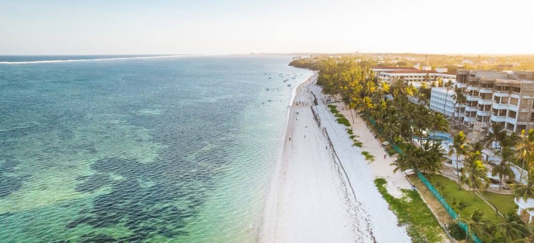 Luftaufnahme vom Diani Beach an der Südküste Kenias mit weißem Sandstrand, türkisfarbenem Wasser und Palmen. Rechts im Bild Hotels, links das ruhige Meer. Die Szene ist in weiches Abendlicht getaucht