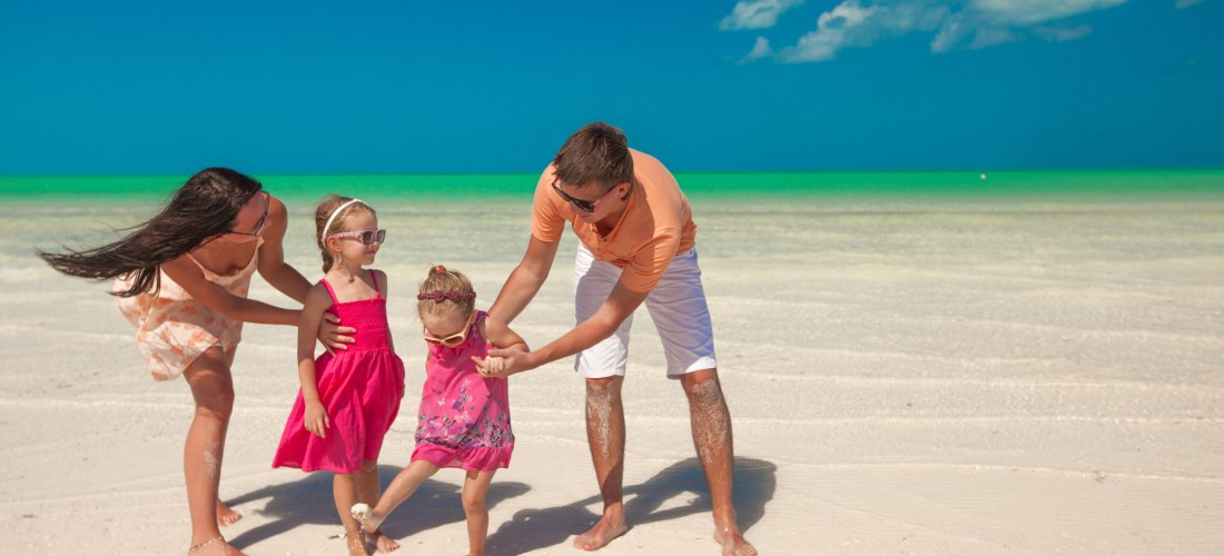 Glückliche Familie mit zwei kleinen Mädchen spielt barfuß am weißen Sandstrand mit türkisfarbenem Wasser unter blauem Himmel