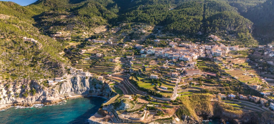 Luftaufnahme einer atemberaubenden mallorquinischen Küstenlandschaft. Im Vordergrund liegt eine felsige Küste mit dramatischen Steilklippen, die ins türkisfarbene Mittelmeer abfallen. Auf den Hügeln dahinter zieht sich ein malerisches Dorf mit hellen Häus