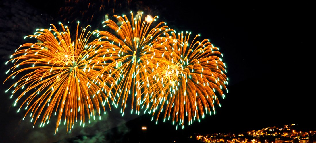 Spektakuläres Feuerwerk über einer Küstenstadt bei Nacht, mit bunten Lichtern am Himmel und reflektierenden Farben im Wasser voller Boote