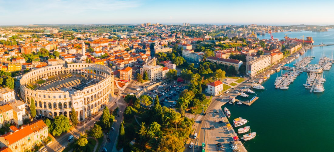 Luftaufnahme der Stadt Pula in Kroatien. Im Vordergrund das gut erhaltene römische Amphitheater, dahinter die Altstadt mit roten Ziegeldächern. Rechts davon ein Jachthafen mit zahlreichen Booten am türkisblauen Meer. Im Hintergrund moderne Stadtviertel un