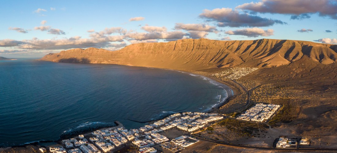 Luftaufnahme von Famara auf Lanzarote mit weißem Dorf, weitem Strand und imposanten Klippen im Hintergrund.