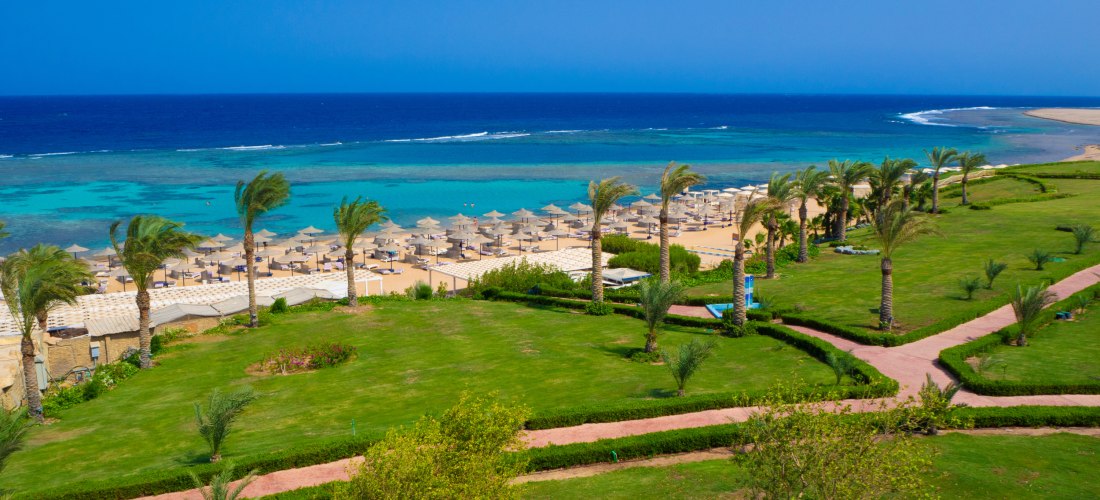Grüne Hotelanlage mit Palmen und Wegen, dahinter Sandstrand mit Sonnenschirmen und türkisfarbenem Meer. Der Himmel ist blau und klar.