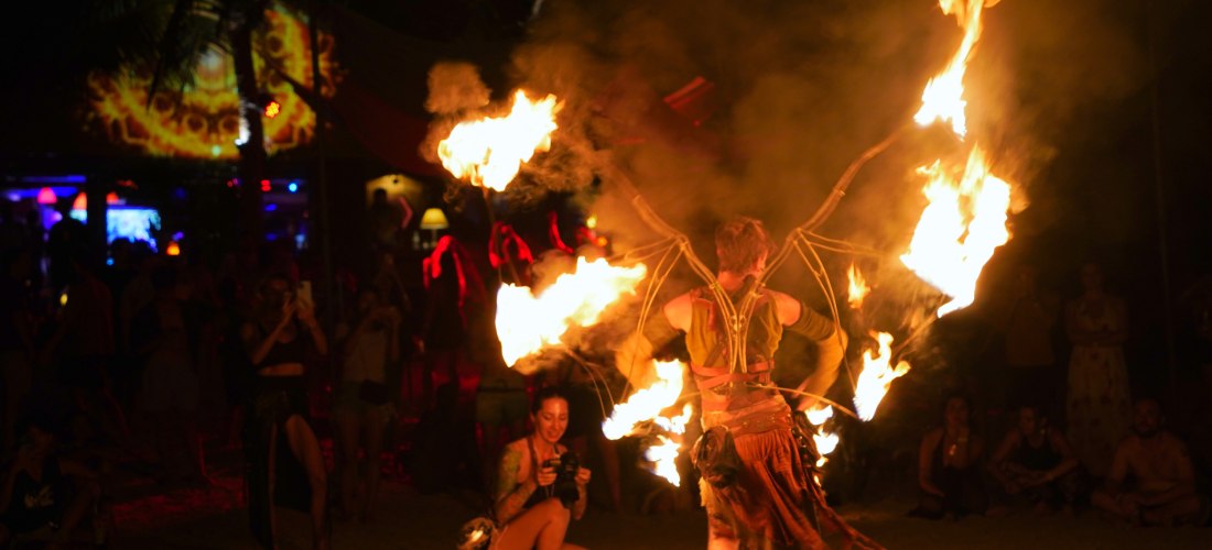 Das Bild zeigt eine faszinierende Feuershow bei Nacht an einem tropischen Strand. Im Vordergrund steht ein Performer in einem kunstvollen Kostüm mit flügelartigen Gestängen, an denen mehrere Flammen brennen. Um ihn herum tanzen Feuerwirbel, während Rauch 