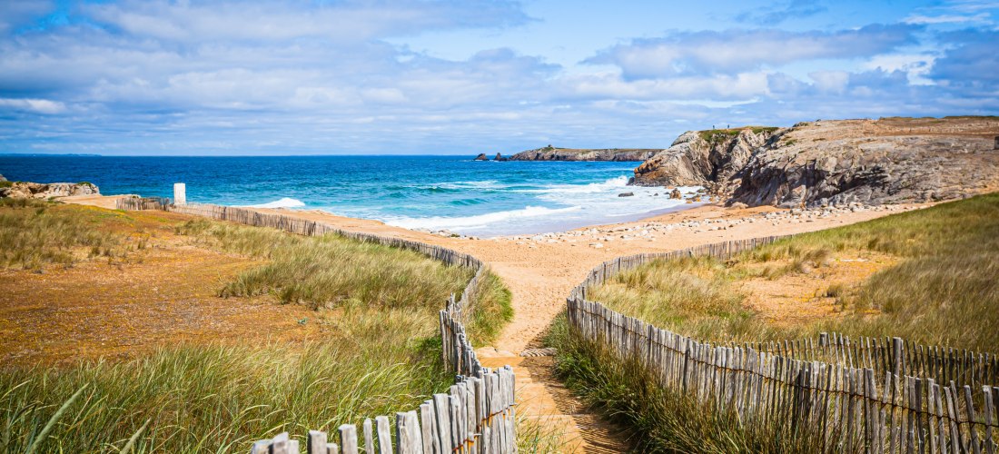 Sandweg zwischen niedrigen Holzlatten-Zäunen führt durch Dünen zum Strand. Rechts Felsen, vorn Dünengras, dahinter türkisblaues Meer mit kleinen Wellen unter teils bewölktem Himmel.