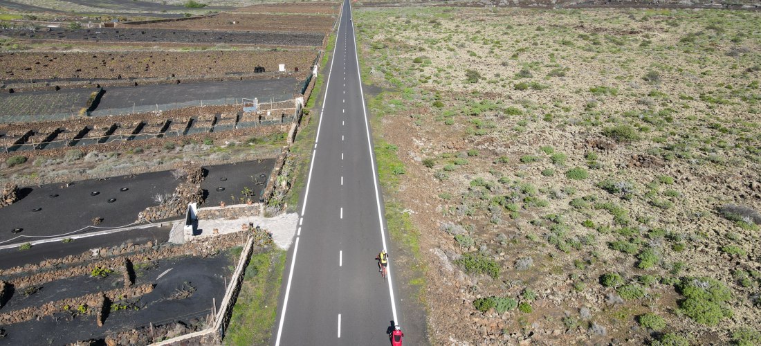 Zwei Radfahrer fahren auf einer langen, geraden Straße durch eine trockene Vulkanlandschaft mit Feldern und niedriger Vegetation. Im Hintergrund erhebt sich ein kegelförmiger Vulkan.