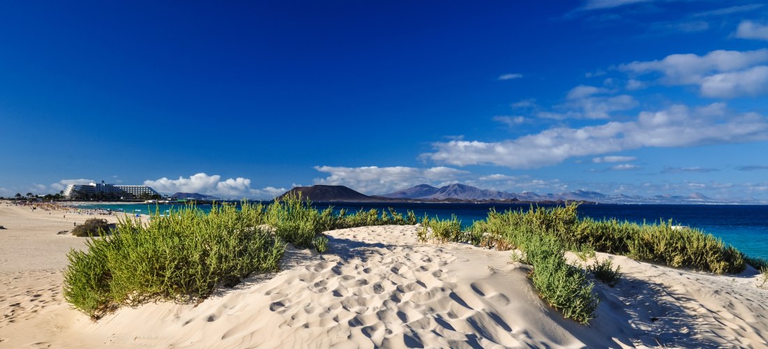 Blick über eine helle Sanddüne mit grüner Vegetation auf einen Strand mit Hotelgebäuden, türkisblauem Meer und einer Vulkanlandschaft im Hintergrund unter tiefblauem Himmel.