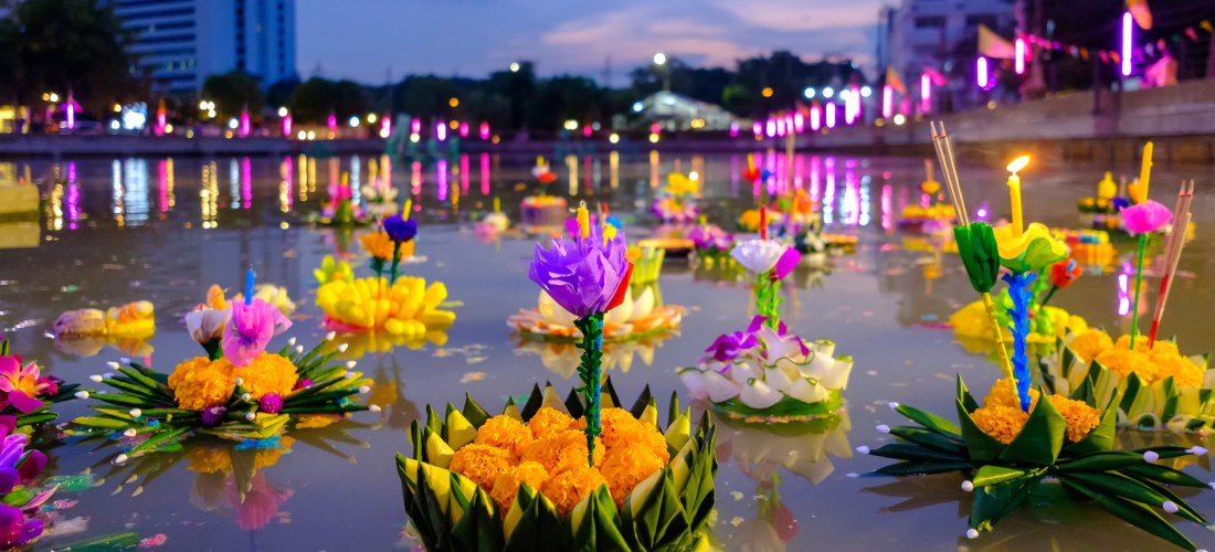 Viele bunte, blumenverzierte Krathongs mit Kerzen und Räucherstäbchen schwimmen auf einem Wasserkanal bei Abenddämmerung. Im Hintergrund leuchten bunte Lichter und Stadthäuser