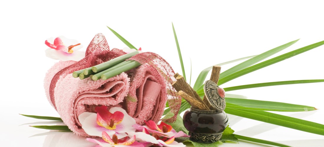 Wellness-Arrangement mit rosa Handtuch, Blumen, Palmblättern, Bambusstäben und einem kleinen Gefäß mit Zimtstangen und orientalischem Deckel. Der Hintergrund ist weiß.