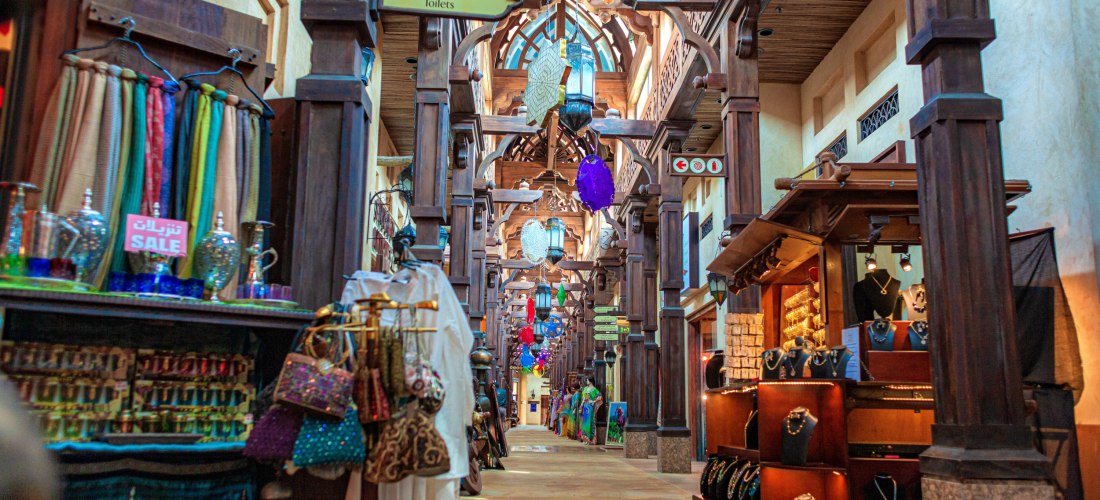 Ein traditionell gestalteter Souk mit Holzsäulen und orientalischer Decke in den Vereinigten Arabischen Emiraten. Stände mit bunten Tüchern, Schmuck, Laternen und Gewürzen säumen den Gang. Das Licht fällt warm durch die Decke und erzeugt eine einladende A