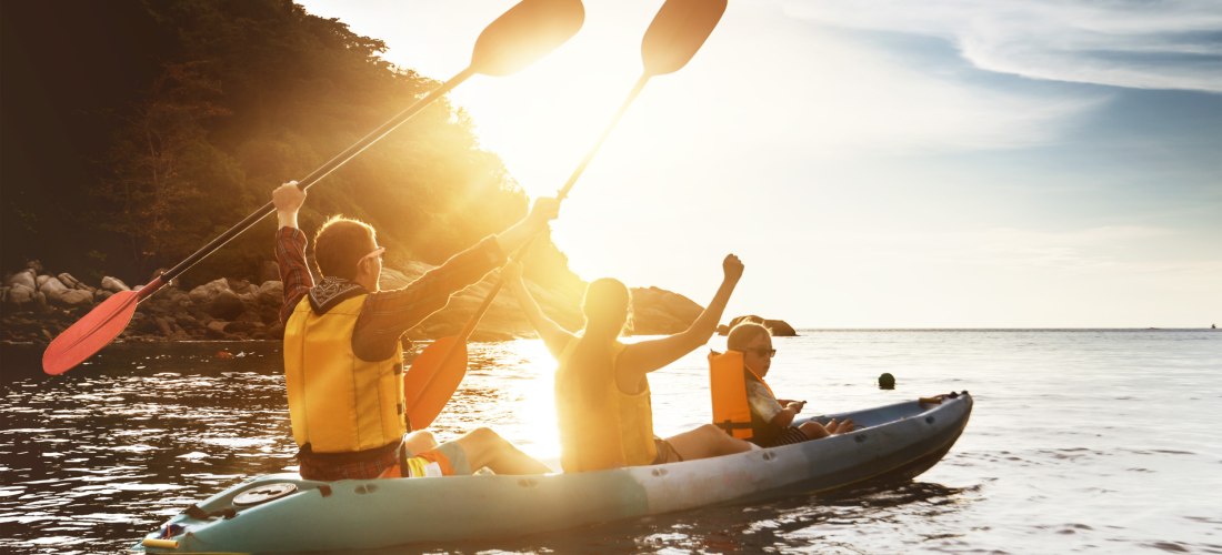 Das Bild zeigt drei Personen – zwei Erwachsene und ein Kind – die gemeinsam in einem Kajak auf dem Meer paddeln. Alle tragen Schwimmwesten, und zwei der Erwachsenen heben ihre Paddel triumphierend in die Luft. Die Szene spielt sich bei Sonnenuntergang ode