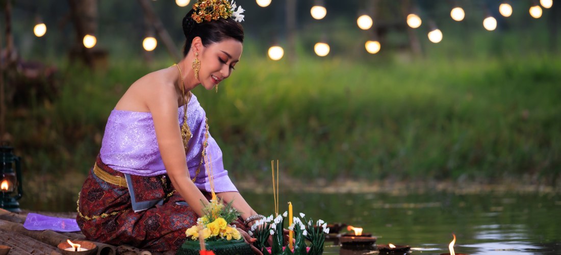 Eine Frau in eleganter thailändischer Kleidung kniet am Ufer eines kleinen Gewässers, um einen mit Blumen, Kerzen und Räucherstäbchen geschmückten Krathong behutsam aufs Wasser zu setzen. Die Szene spielt bei Nacht, im Hintergrund sind warme Lichtpunkte d