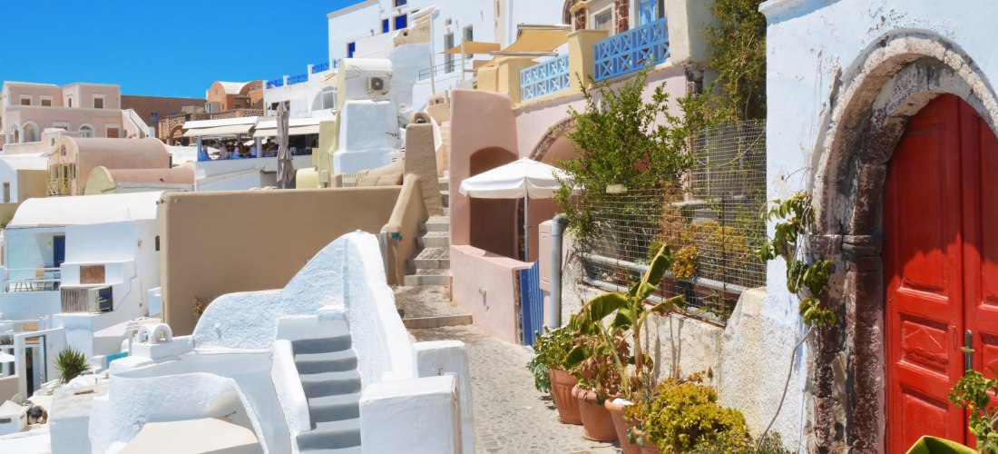 Ein schmaler Weg mit kleinen Treppenstufen windet sich durch eine Häuserreihe auf Santorini. Die Gebäude sind in hellen Farben gestrichen, hauptsächlich Weiß mit Akzenten in Blau, Rosa und Beige. Rechts steht eine markante rote Tür in einem alten gemauert