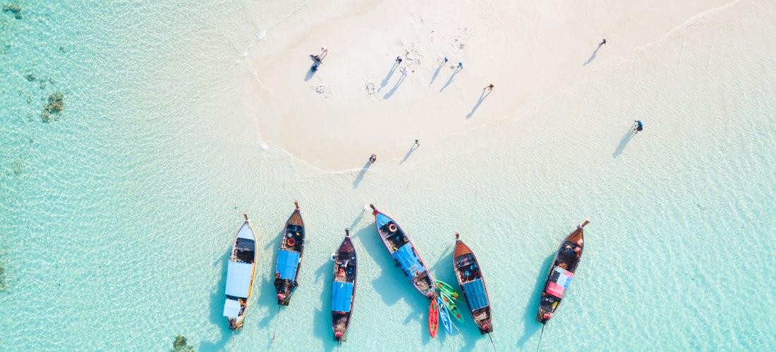 Drohnenaufnahme von sieben traditionellen Longtail-Booten, die nebeneinander im türkisfarbenen, klaren Wasser einer Sandbank auf Phuket liegen. Einige Menschen spazieren barfuß über den hellen Sand.