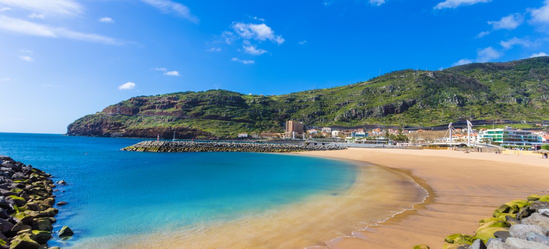 Breiter Sandstrand Praia da Calheta auf der Insel Madeira, umgeben von türkisblauem Meer und einer geschützten Bucht. Im Hintergrund erheben sich grüne Hügel mit kleinen Gebäuden und einer Promenade, unter einem klaren, blauen Himmel.