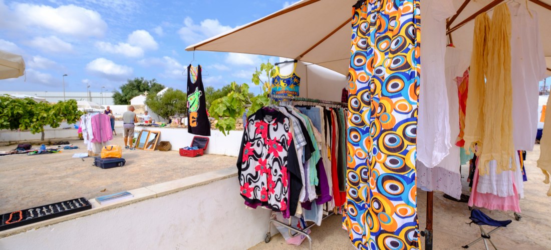 Offener Marktstand unter einem großen Sonnenschirm, an dem bunte Sommerkleidung auf Kleiderständern hängt. Im Vordergrund auffällige Muster in Orange, Blau und Weiß. Im Hintergrund weitere Stände und Besucher.