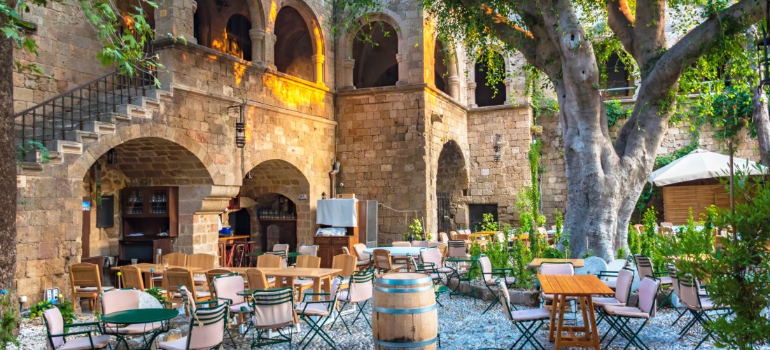 Das Bild zeigt einen idyllischen Innenhof in der Altstadt von Rhodos, der von einer mittelalterlichen Steinarchitektur geprägt ist. Massive Steinbögen und Arkaden umrahmen den Platz, auf dem zahlreiche Tische und Stühle stehen. Einige der Tische sind rund