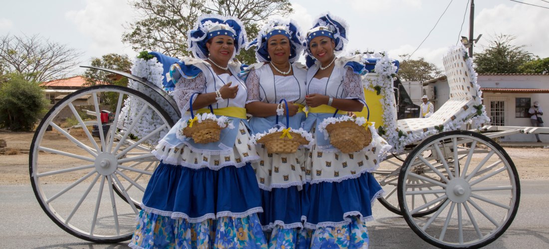 Drei Frauen in farbenfroher traditioneller Kleidung stehen nebeneinander vor einer dekorierten weißen Kutsche mit großen Rädern. Sie tragen blau-weiße Kleider mit Spitzenapplikationen, gelben Schärpen und passenden Hauben. Jede hält ein kleines Körbchen i