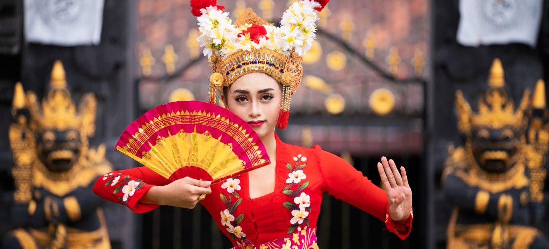 Eine balinesische Tänzerin in traditioneller roter Kleidung mit aufwendigem goldverziertem Kopfschmuck und Blütenschmuck führt einen klassischen Tanz auf. In der einen Hand hält sie einen roten Fächer, die andere Hand ist in einer präzisen Geste erhoben. 