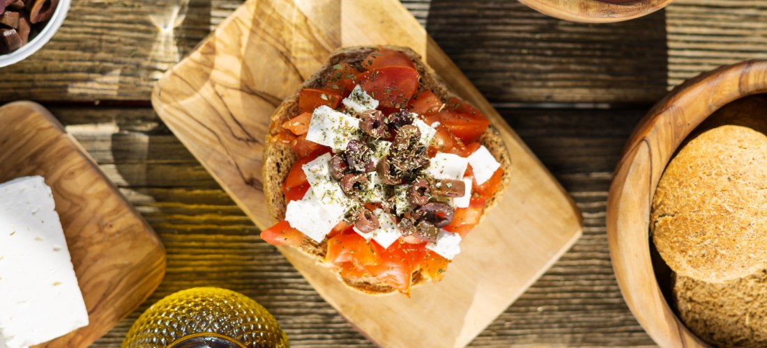 Ein traditioneller griechischer Dakos auf einem Holzbrett: Geröstetes Gerstenbrot belegt mit gewürfelten Tomaten, Fetakäse, Oliven und Kräutern. Umgeben von weiteren Zutaten wie Oliven, Tomaten, Brot, Feta und einer Flasche Olivenöl auf einem rustikalen H