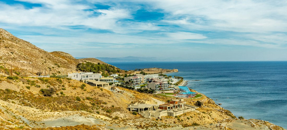 Blick auf ein modernes Hotelresort an einer felsigen Küste mit direktem Zugang zum Meer. Das Resort liegt eingebettet zwischen trockenen Hügeln mit spärlicher Vegetation. Im Vordergrund sind unfertige Gebäude oder Baustellen zu erkennen. Der Himmel ist le