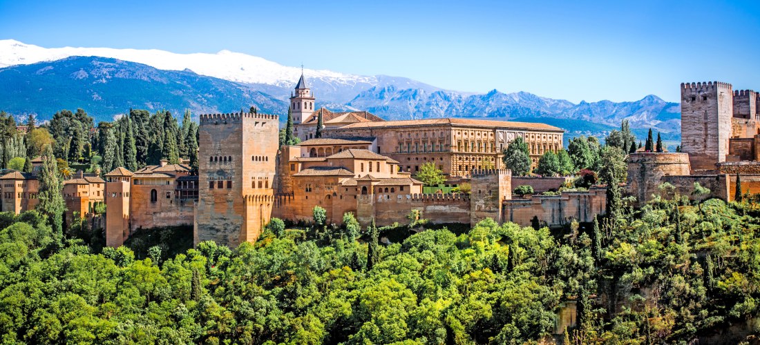 Die Alhambra in Granada mit ihren rötlich-braunen Türmen vor schneebedeckten Bergen der Sierra Nevada, umgeben von grünen Bäumen.