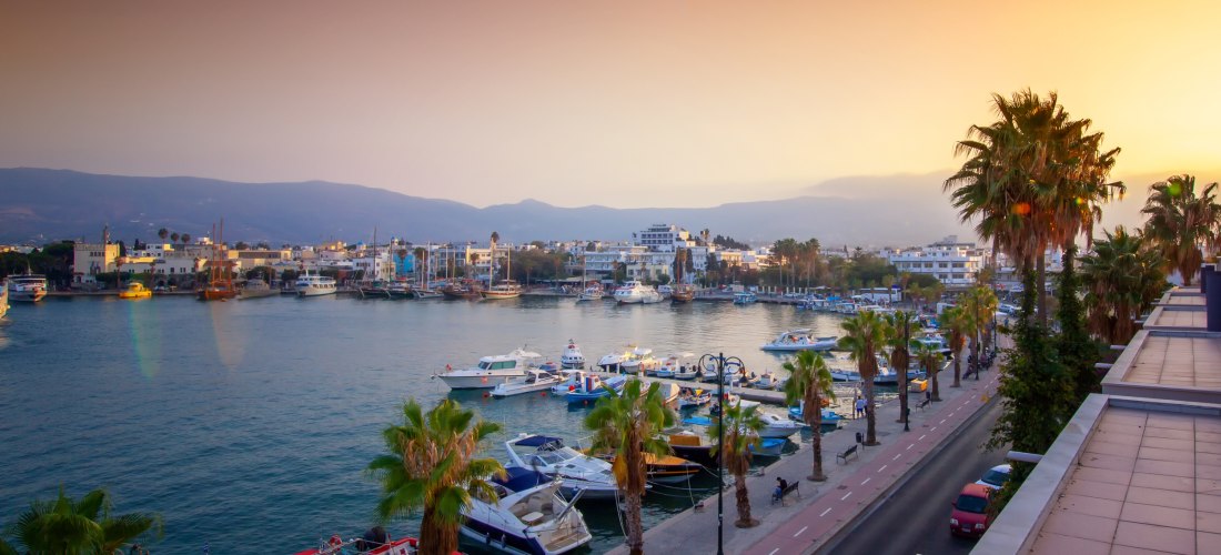 Blick auf den Yachthafen von Kos-Stadt bei Sonnenuntergang. Die Promenade ist gesäumt von Palmen, und zahlreiche Boote liegen im ruhigen Wasser. Im Hintergrund leuchten die Gebäude der Stadt vor einer Bergsilhouette in warmem Licht.