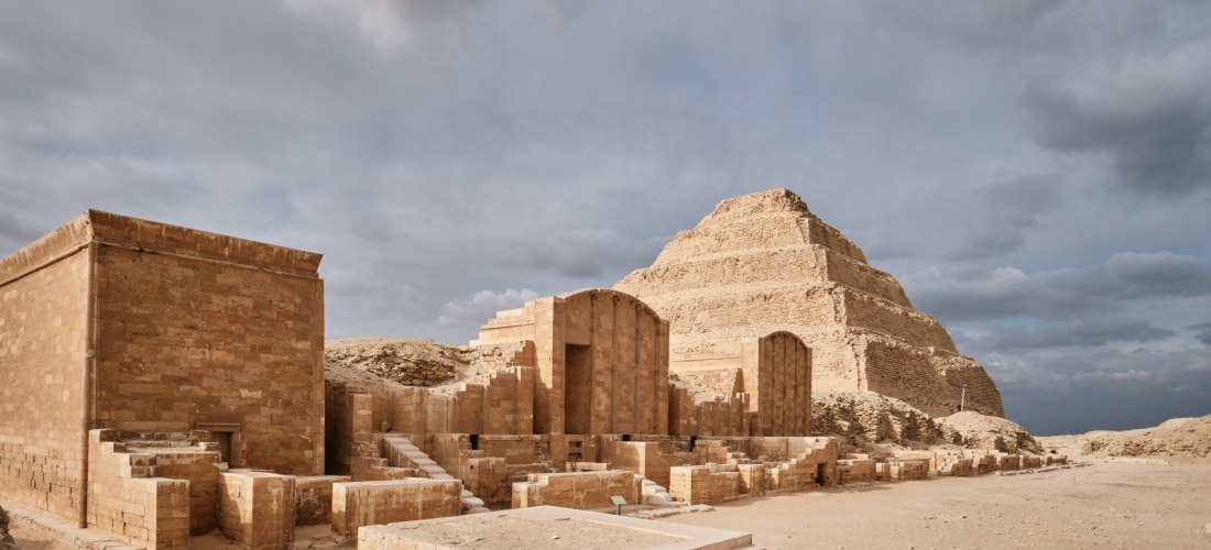 Blick auf die Stufenpyramide von Sakkara in Ägypten, eine der ältesten Pyramiden der Welt. Im Vordergrund sind gut erhaltene Überreste antiker Tempel- und Grabarchitektur zu sehen, bestehend aus großen Sandsteinblöcken. Die Pyramide erhebt sich im Hinterg