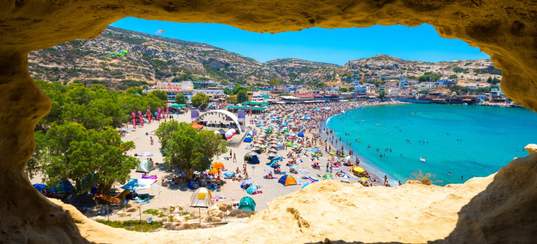 Blick aus einer natürlichen Felsengrotte auf den belebten Strand von Matala, Kreta. Der Sandstrand ist voller bunter Sonnenschirme, Badetücher und Menschen. Am Rand befindet sich eine Bühne mit Festivalcharakter. Dahinter erstreckt sich eine Bucht mit tür