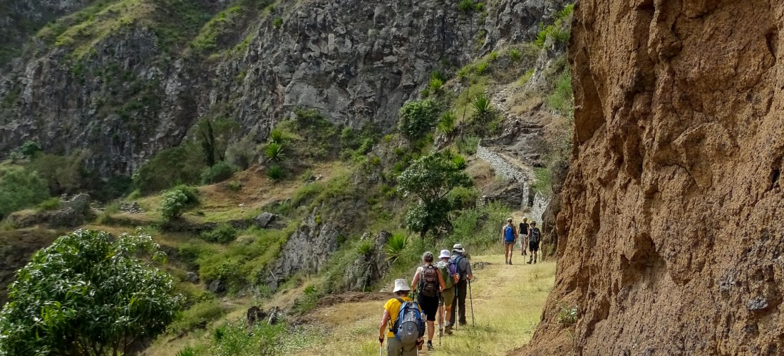 Wandergruppe mit Rucksäcken und Wanderstöcken auf einem schmalen Pfad entlang einer Felswand in einer bergigen, grün bewachsenen Landschaft. Die Wandernden bewegen sich durch eine tiefe Schlucht, umgeben von steilen Hängen.