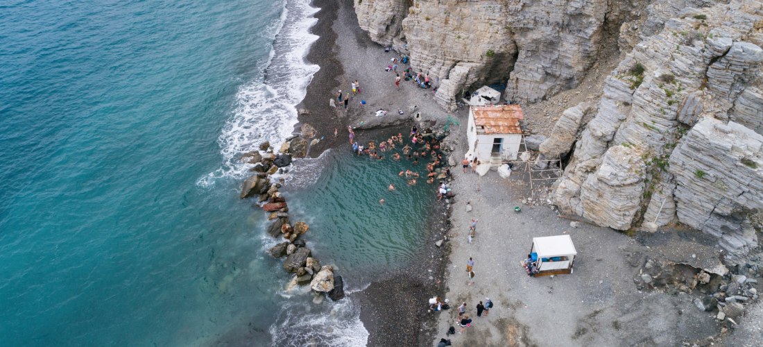  Dieses Bild zeigt eine kleine Bucht an einer felsigen Küste. In der Mitte befindet sich ein natürliches Becken mit warmem Wasser, das von Steinen vom offenen Meer abgegrenzt ist. Darin baden etwa 20 bis 30 Menschen. Der schmale Strand besteht aus dunklem