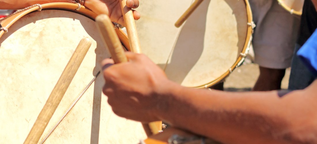 Mehrere Menschen spielen traditionelle Trommeln mit Holzstöcken bei einem Straßenfest. Die Szene zeigt Nahaufnahmen von Händen, Trommeln und rhythmischer Bewegung. Die Kleidung ist sommerlich und farbenfroh.