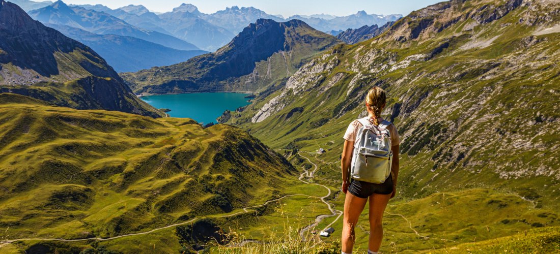 Panoramablick über grüne Alpentäler mit türkisblauem Bergsee; Wanderin mit Rucksack steht am Grat bei klarem Sommerwetter.