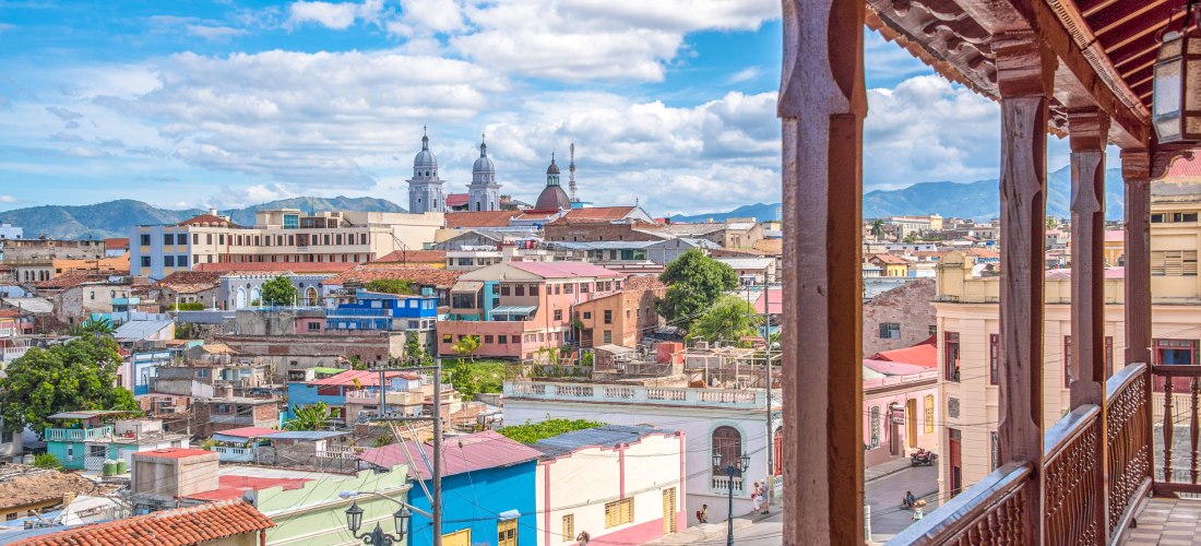 Santiago de Cuba – Blick auf die historische Altstadt mit Kathedrale Panoramablick von einem Holzbalkon auf die farbenfrohen Häuser der Altstadt von Santiago de Cuba mit der imposanten Kathedrale im Hintergrund und den Bergen der Sierra Maestra.