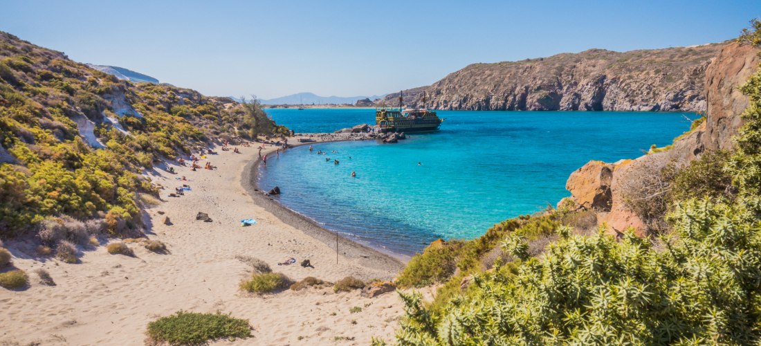 Kleine geschützte Badebucht mit goldenem Sandstrand und türkisfarbenem Wasser auf Kos. Links sonnen sich Menschen am Strand, einige schwimmen im Wasser. Rechts liegt ein traditionell gestaltetes Ausflugsboot vor Anker. Umgeben ist die Bucht von Felsen und