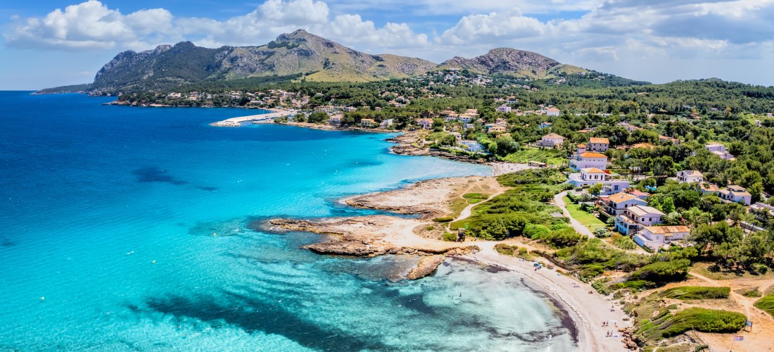 Luftaufnahme der Küste von Nordmallorca mit türkisblauem Meer, Sandstrand, Felsen und Blick auf das Tramuntana-Gebirge bei Alcúdia