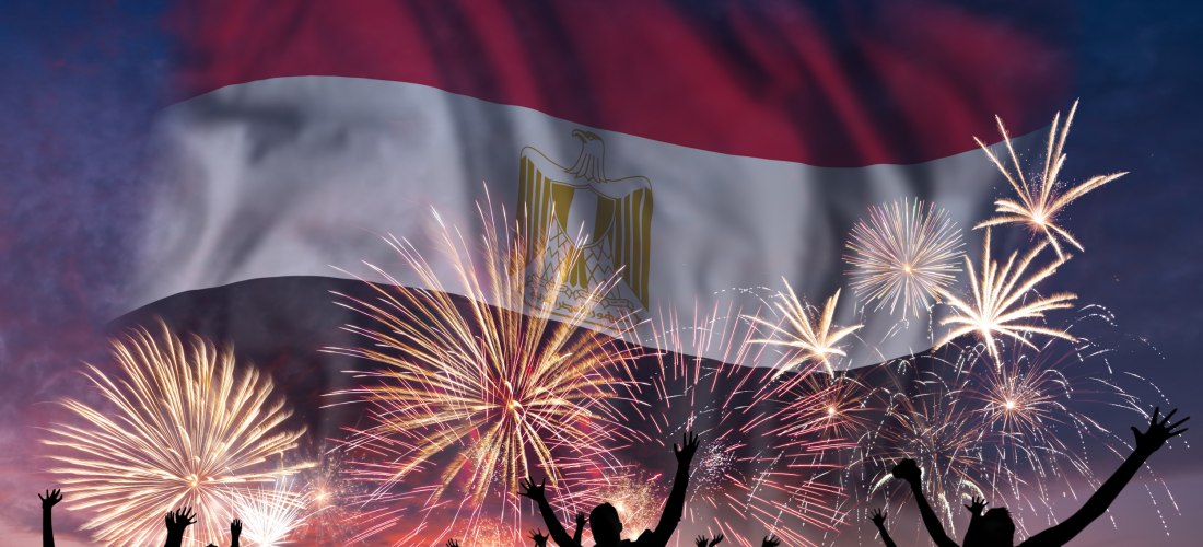 Eine Menschenmenge feiert ausgelassen mit erhobenen Armen vor einem Feuerwerk am Abendhimmel. Im Hintergrund ist groß die ägyptische Flagge eingeblendet.