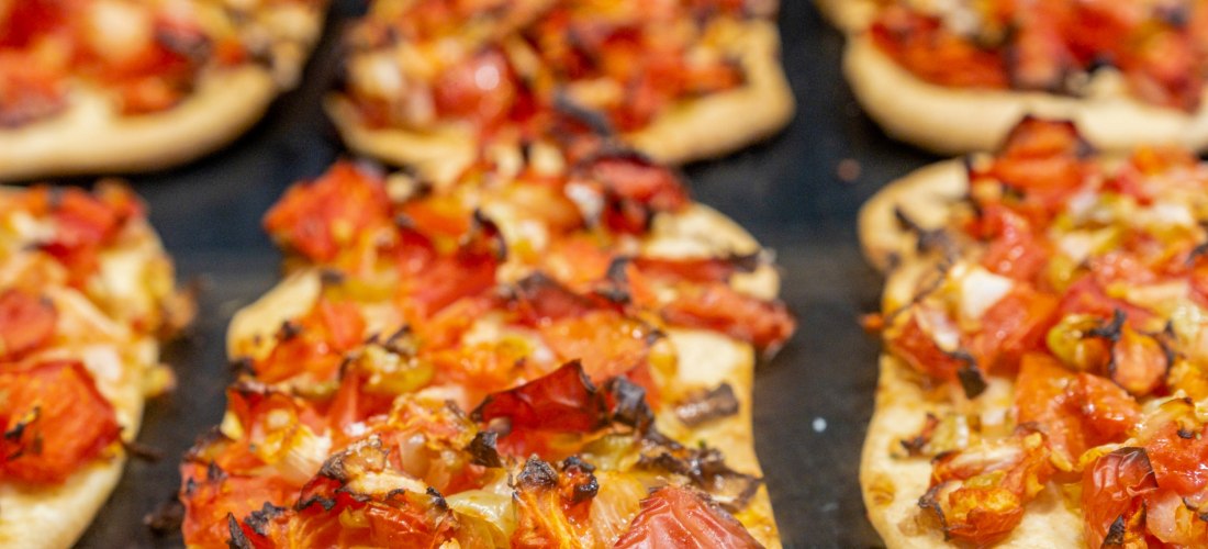 Mehrere Portionen einer typischen mallorquinischen Coca – ein herzhaftes Fladenbrot – belegt mit gehackten Tomaten, Zwiebeln und Paprika. Die Zutaten sind leicht angeröstet, der Teig goldbraun gebacken. Die Coca liegt auf einem dunklen Blech und wirkt fri