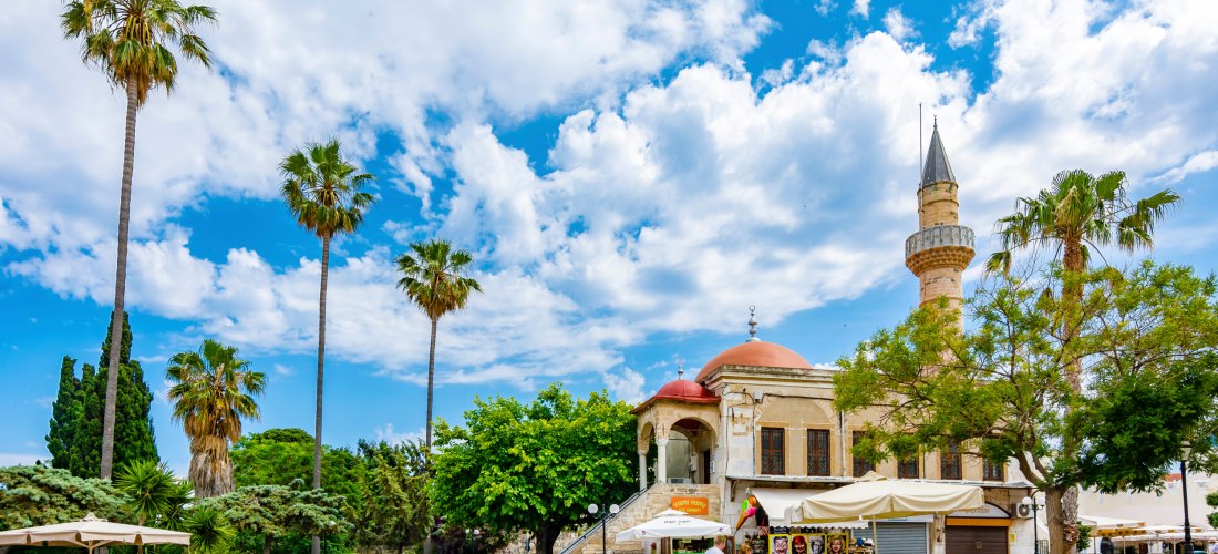 Lebhafter Platz in der Stadt Kos mit Palmen, einem Verkaufsstand und dem historischen Gebäude der Defterdar-Moschee mit roter Kuppel und Minarett. Menschen bummeln gemütlich unter blauem Himmel mit dekorativen Wolken.