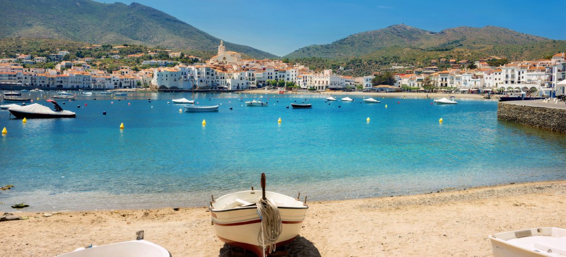 Blick von einem Sandstrand mit kleinen Booten auf das klare blaue Meer und die malerische Küstenstadt Cadaqués in Katalonien, Spanien. Im Hintergrund steigen weiße Häuser an einem Hügel auf, überragt von einer Kirche mit hellem Turm.
