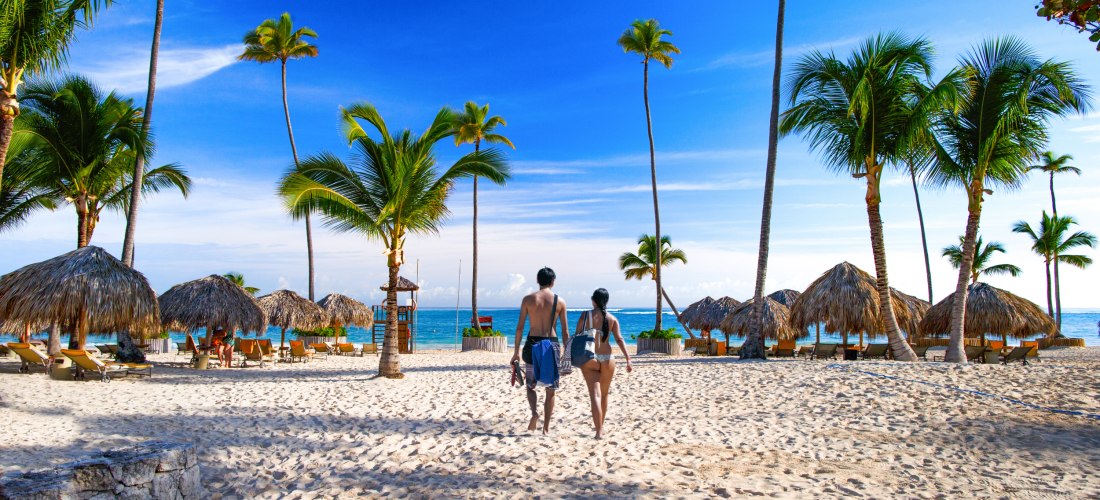 Paar in Badesachen spaziert auf einem tropischen Sandstrand mit Palmen und Strohhütten, Blick auf das türkisfarbene Meer unter blauem Himmel