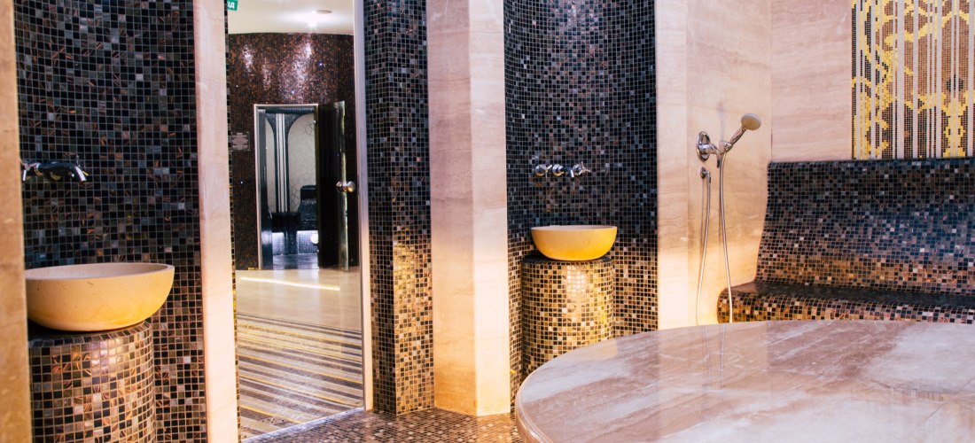 Modern gestalteter Hamam-Raum mit dunklem Mosaik, zwei Waschbecken, einer geschwungenen Sitzbank, Handbrause und einem großen beheizten Marmortisch in der Mitte. Sanfte Beleuchtung sorgt für eine entspannte Atmosphäre.