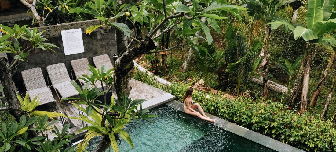 Eine Frau sitzt entspannt am Rand eines privaten Pools inmitten tropischer Vegetation auf Bali. Umgeben von Palmen, Bananenpflanzen und dichtem Grün wirkt der Poolbereich ruhig und abgeschieden. Neben dem Pool stehen mehrere leere Sonnenliegen auf einer H