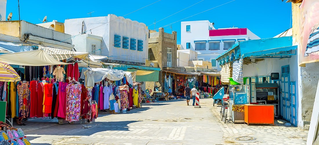 Eine bunte Souk-Straße in Tunesien mit zahlreichen Ständen und Geschäften. Bunte Stoffe und Gewänder hängen an Kleiderstangen, während Händler ihre Waren präsentieren. Die Szene ist lebendig, voller Farben und spiegelt das pulsierende Leben der Medina wid