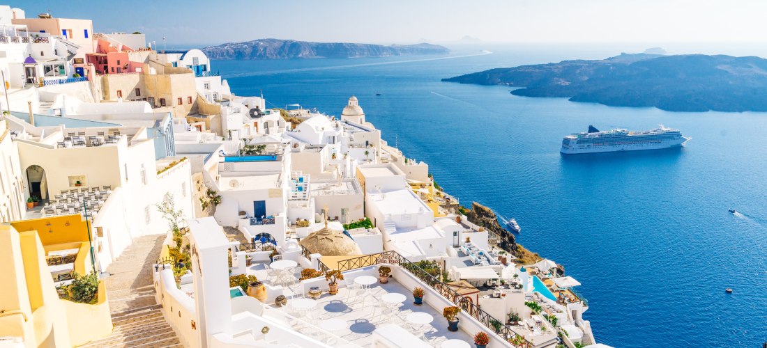 Fira auf Santorini: weiße Kykladenhäuser und Terrassen über der Caldera, unten ein Kreuzfahrtschiff im tiefblauen Meer