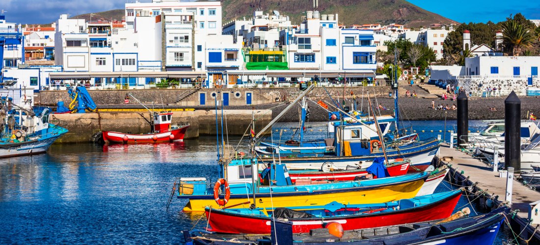 Bunte Fischerboote im Hafen von Agaete auf Gran Canaria vor weiß-blauen Häusern und grünen Bergen im Hintergrund.