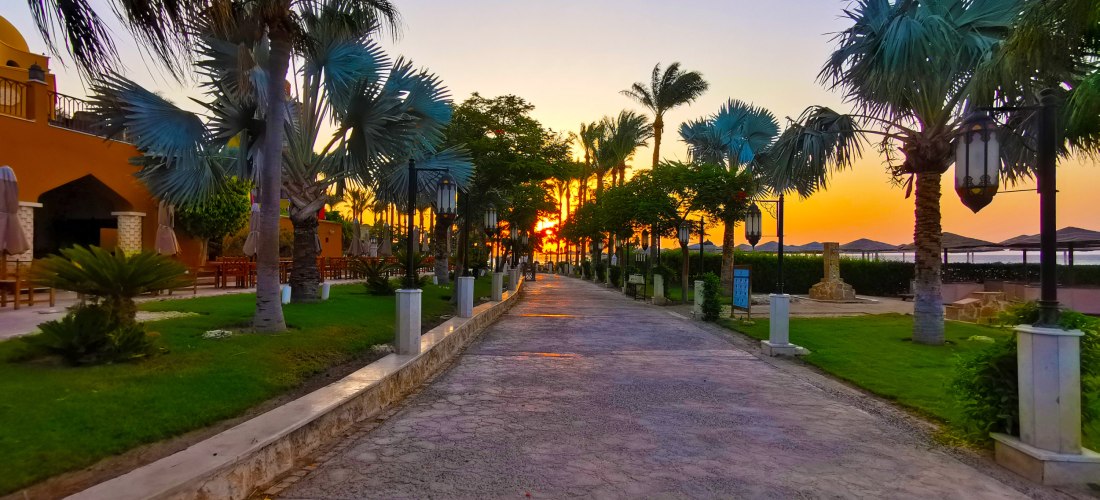 Sonnenuntergang auf einer palmengesäumten Promenade einer Hotelanlage, mit Blick auf das Meer. Rechts und links sind Laternen, Palmen und mediterrane Gebäude zu sehen.