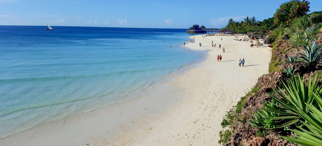 Strand auf Sansibar mit weißem Sand, türkisblauem Meer und traditionellem Dhow-Segelboot im Hintergrund