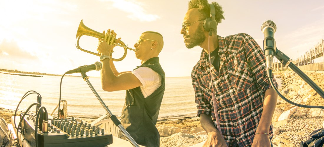 Zwei Musiker am Strand bei Sonnenuntergang. Links spielt ein Mann Trompete vor einem Mikrofon, rechts steht ein DJ an einem Mischpult mit Kopfhörern. Die goldene Sonne reflektiert auf dem Wasser, im Hintergrund eine ruhige Strandpromenade