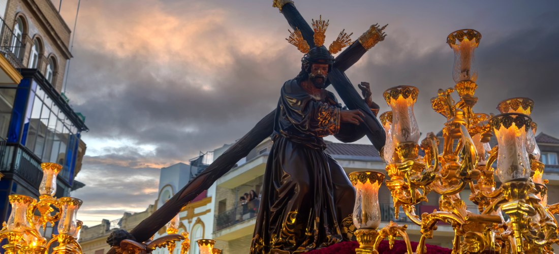 Eine große, kunstvoll geschmückte Jesusstatue mit Kreuz wird bei einer religiösen Prozession durch die Straßen getragen. Die Figur zeigt Jesus mit Dornenkrone und goldenen Strahlen um den Kopf, bekleidet mit einem dunklen Gewand. Die Plattform ist mit gol